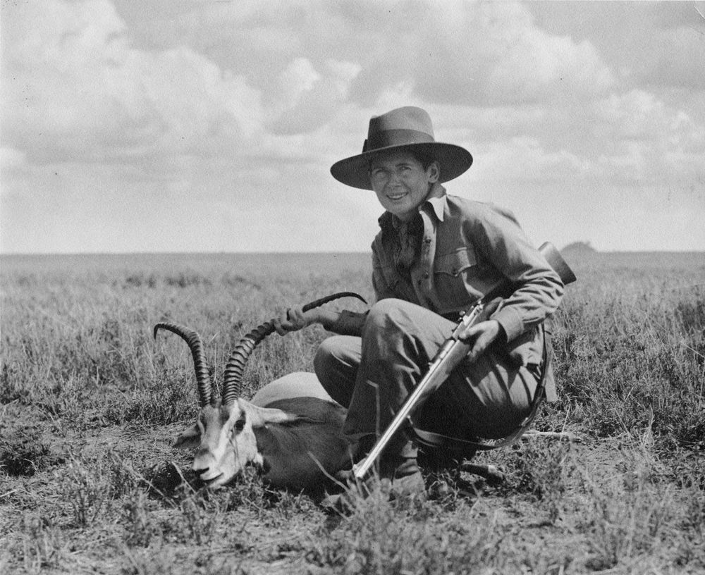 A black and white image of Pauline Pfeiffer Hemingway on safari in Africa.  She is wearing hunting garb and a wide-brimmed hat.  She kneels, holding a rifle, posing by the body of a gazelle.