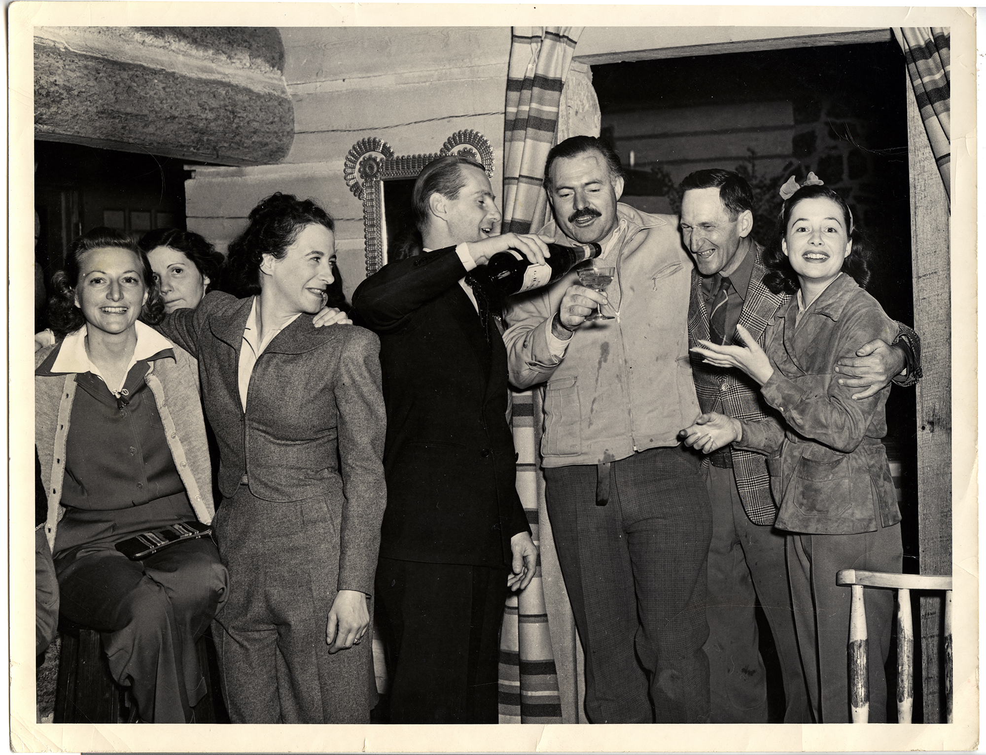 A black and white photograph, slightly sepia with age, showing Ernest Hemingway and six other people (four women; two men) at a party in a log cabin.  All are dressed casually save one man who is pouring champagne into Hemingway's glass; he wears a dark suit, white shirt, and tie.